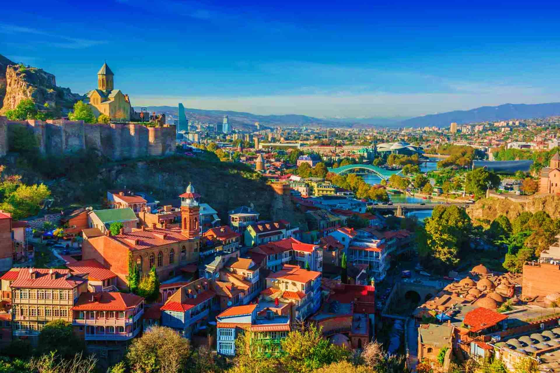 Georgia Visa Aerial view of Tbilisi, Georgia showing the Metekhi Church on cliffs, Mtkvari River, and cityscape with mountains in backgrou